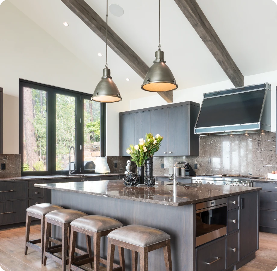 Spacious kitchen with dark wood cabinets, large marble island with stools, stainless steel appliances, pendant lights, and a view of trees through large windows.