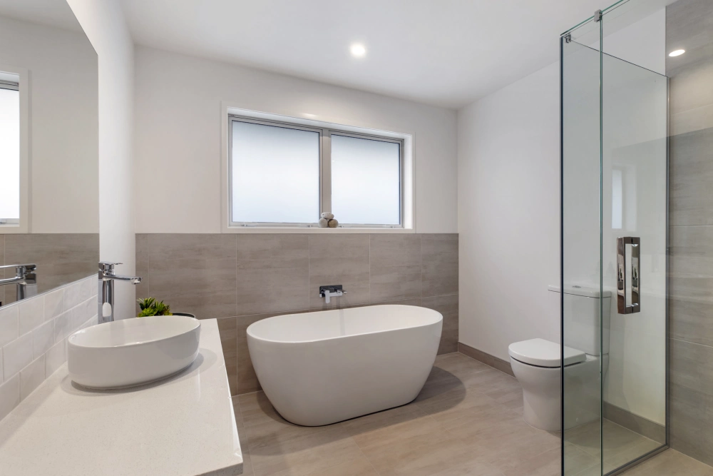 Modern bathroom with a freestanding bathtub, glass-enclosed shower, oval sink, and toilet. Minimalist design with neutral tones and a large window with frosted glass.