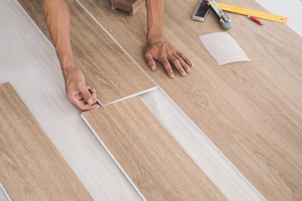 Hands are installing wood-style laminate flooring, aligning the planks on a white underlayment. Tools, such as a pencil and tape measure, are scattered nearby.