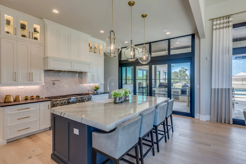 Modern kitchen with a large island, marble countertops, pendant lights, and a gas stove. Glass doors lead to an outdoor area.