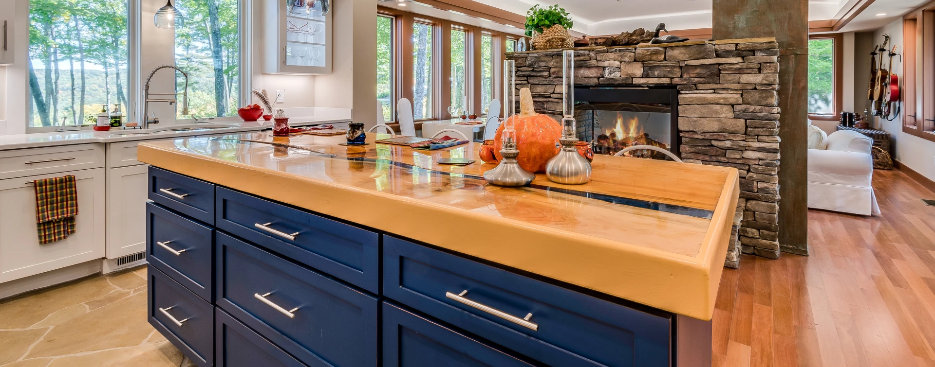 A modern kitchen with a blue island countertop, wooden floors, and a stone fireplace. Large windows provide natural light. Various decorative items are displayed on the island.