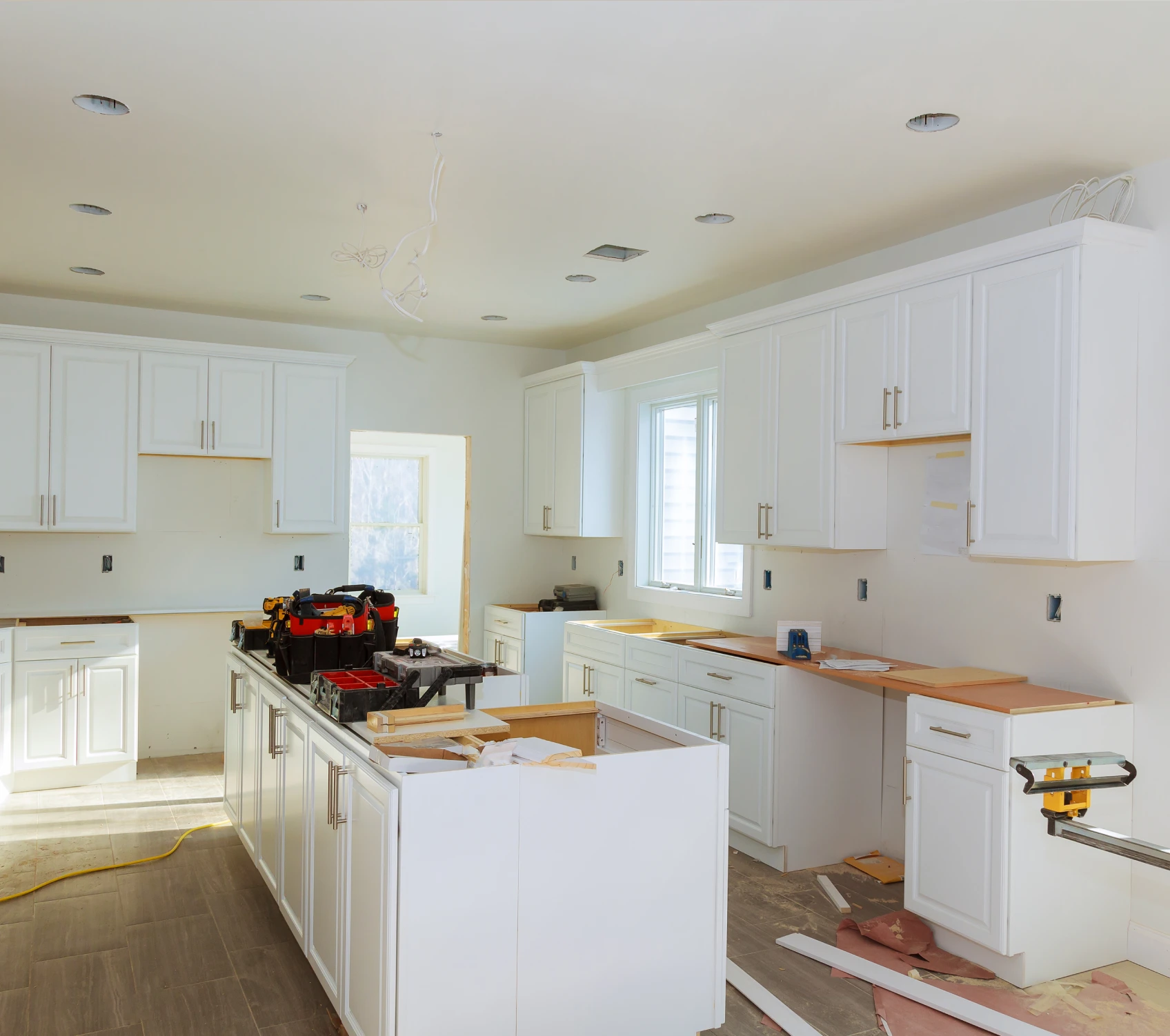 A kitchen under construction with white cabinets, tools on the island, and several open drawers. A window is on the far wall, and materials are scattered around the floor.